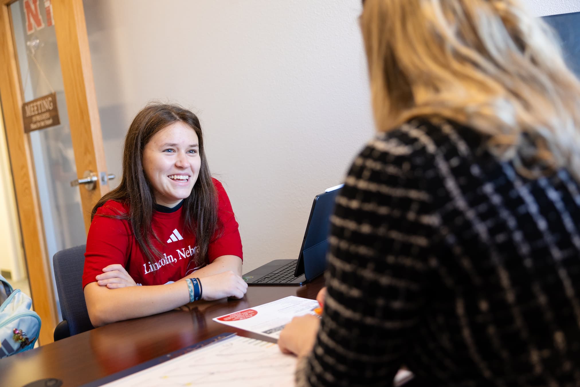 Woman in a red shirt smiling during a conversation at a wooden desk.