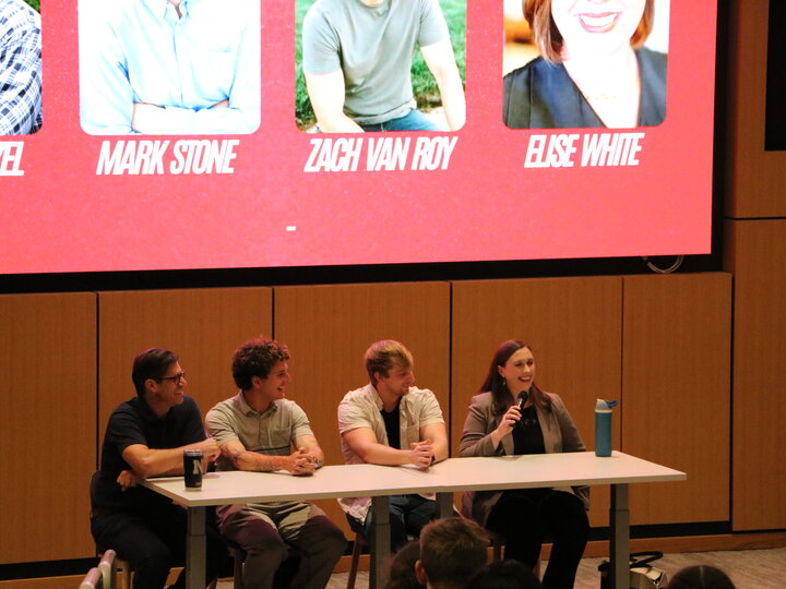 Four panelists seated at a table speaking to an audience, with a red presentation screen behind them.