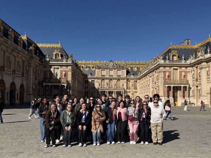 A large group of 25 students gathers in a group photo in front of a grand, stone building with a dark roof that forms a square around them in Paris.
