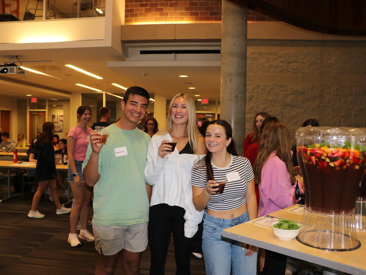 Three people smiling and holding drinks in a bright indoor event space with a fruit punch dispenser nearby.