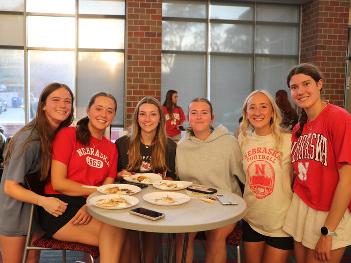 Group of six smiling young women sitting around a table with plates of food, wearing Nebraska shirts.