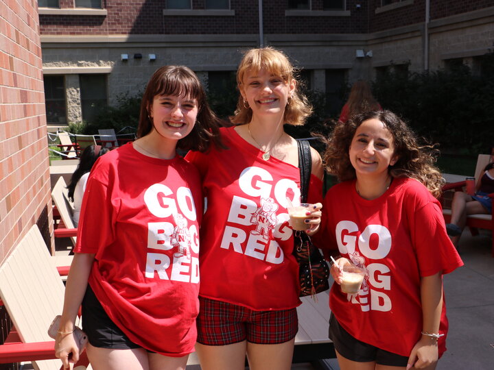 Three smiling women wearing red "Go Big Red" shirts, standing outdoors in a sunny courtyard.