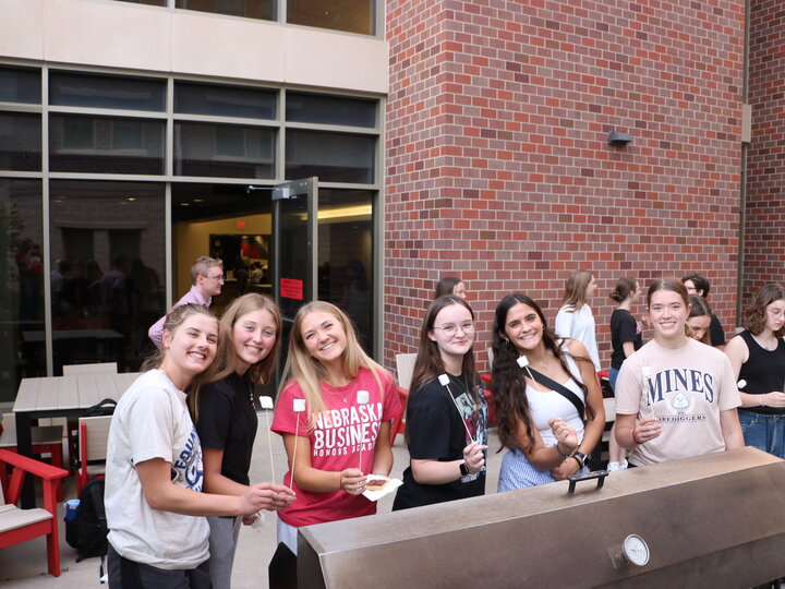 Group of six smiling young adults roasting marshmallows at an outdoor event near a brick building.