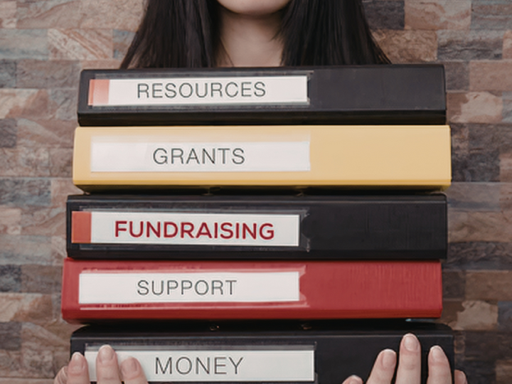 Person holding stacked binders labeled Resources, Grants, Fundraising, Support, and Money.