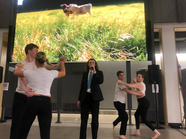 Dancers move around a person singing in the center with an image of a field projected behind them.