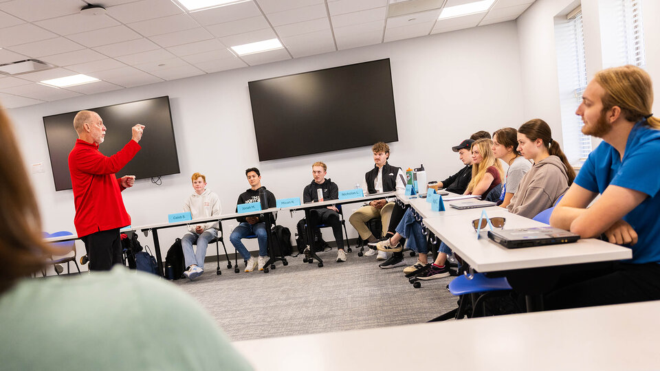 An older white man in a red hoodie and black pants speaks to a class of college students.